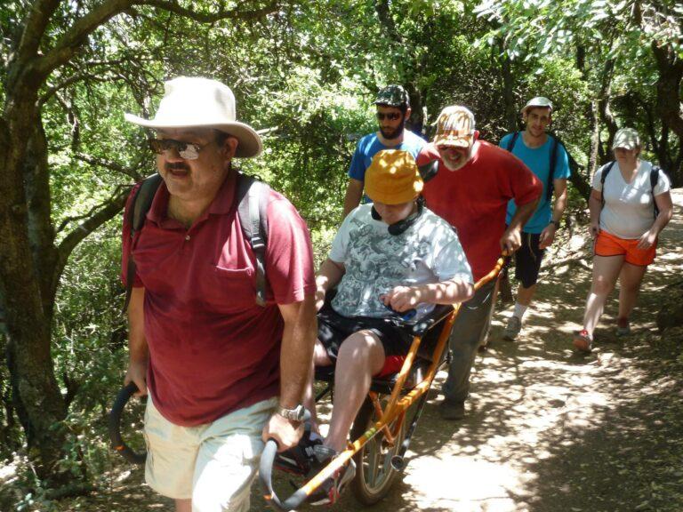 Two men assisting a traveler in an all-terrain wheelchair on a hike together with other hikers.