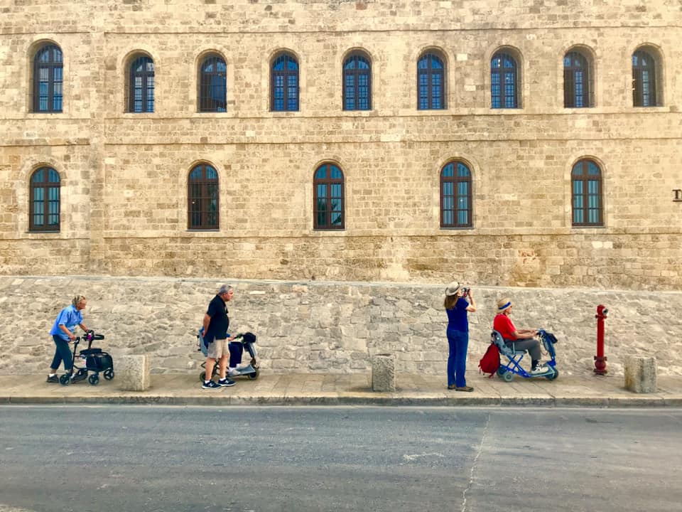 A group of travelers, some of whom are using mobility scooters, in front of an old brick building in Israel.