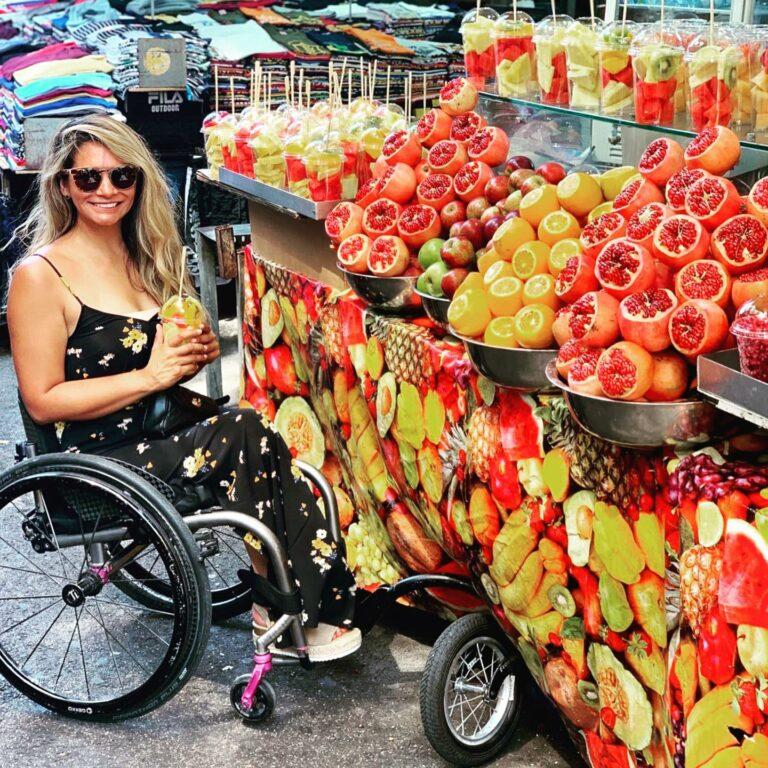 A female traveler in a flowery dress and a wheelchair smiling whilst holding a fruit salad from a colorful fruit stand in the street in Israel.